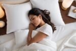 Woman with long dark hair soundly asleep in bed with white sheets pillows and blankets